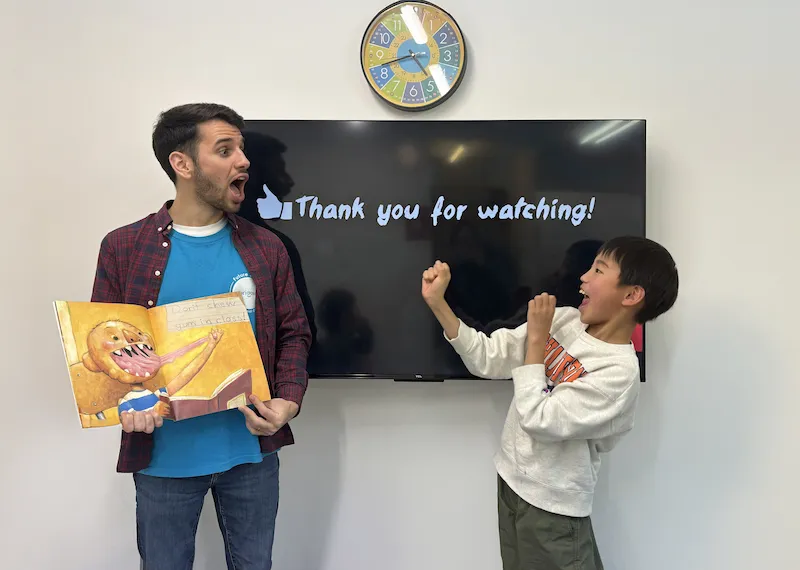 キュリオキッズの外国人講師と子供がテレビの前で本を読んでいる様子