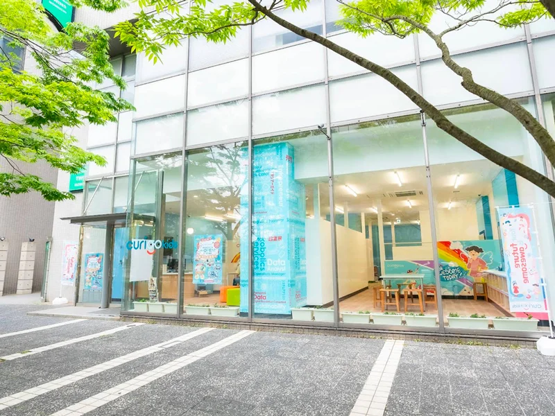 緑の木々に囲まれたガラス張りの現代的な店舗外観のCuriooKids百道浜スタジオ